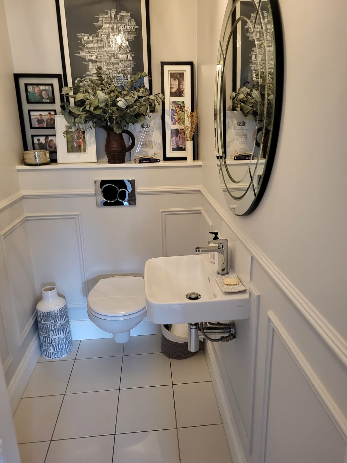 Elegant bathroom with white paneling, decorative mirror, shelving with family photos and plants