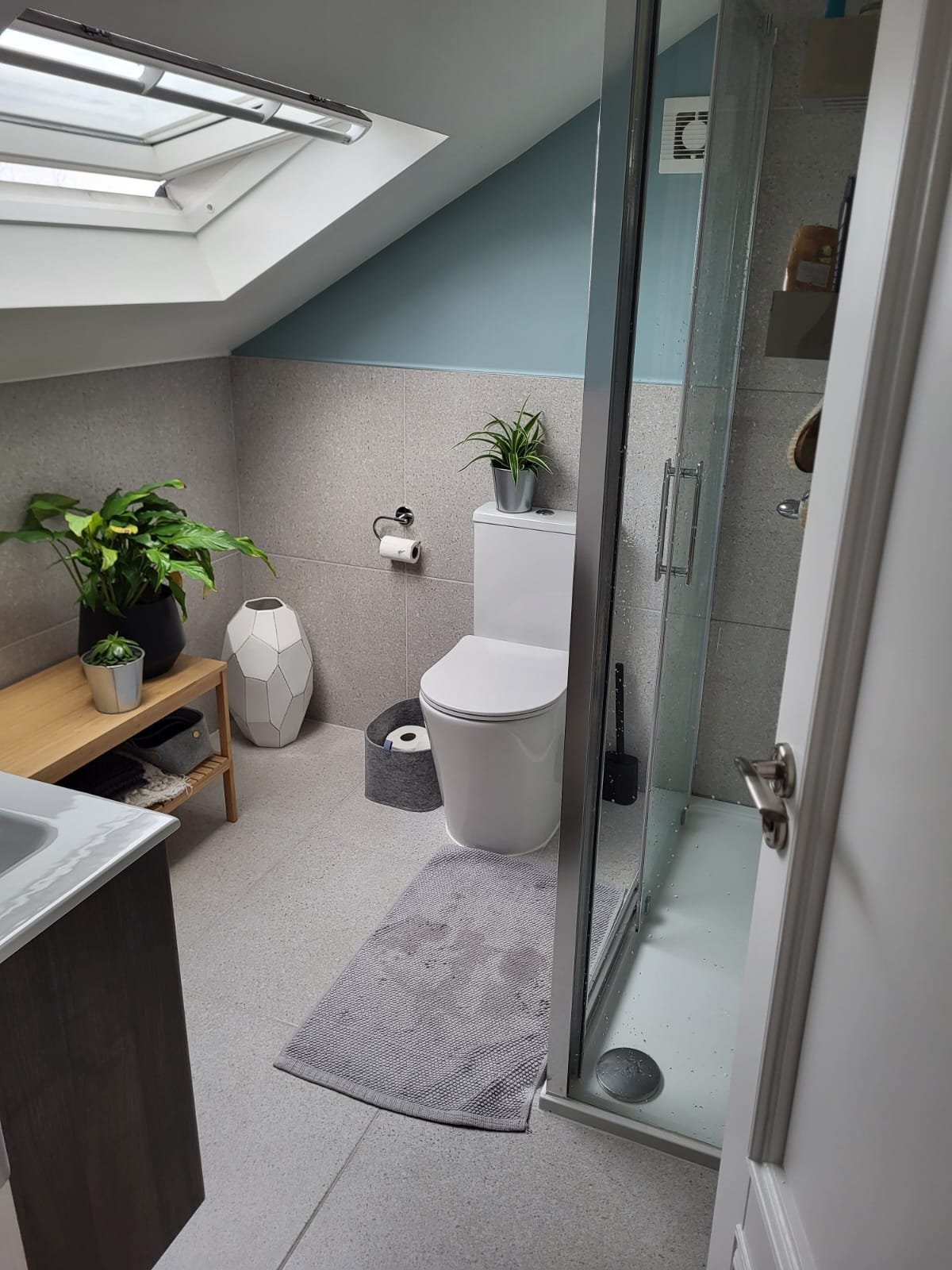 Modern ensuite bathroom with gray tile, white toilet, wooden shelf, and glass shower enclosure