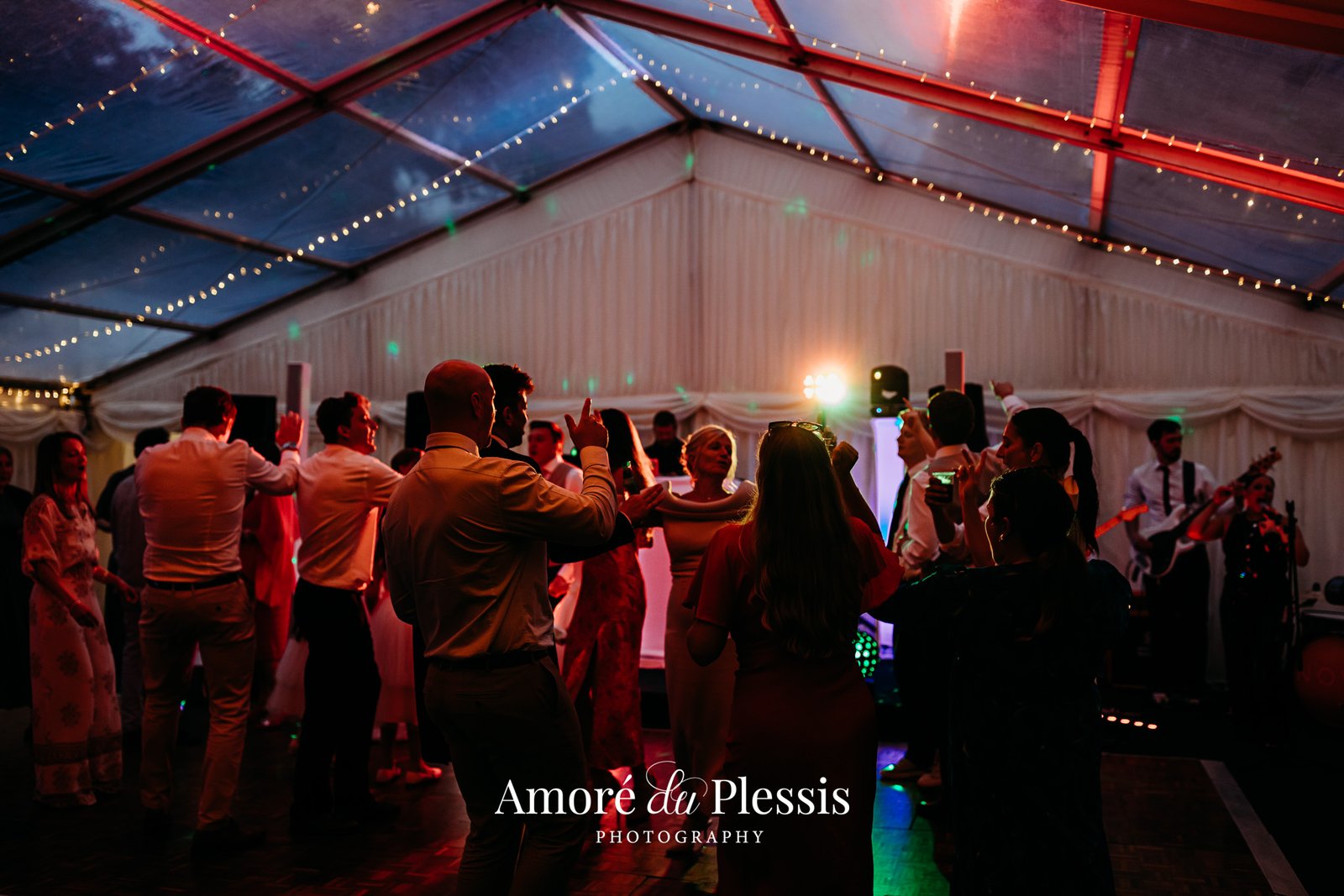 Elegant tent event venue with red lighting, guests dancing, live band performing, decorated ceiling with string lights and red draping, professional event photography