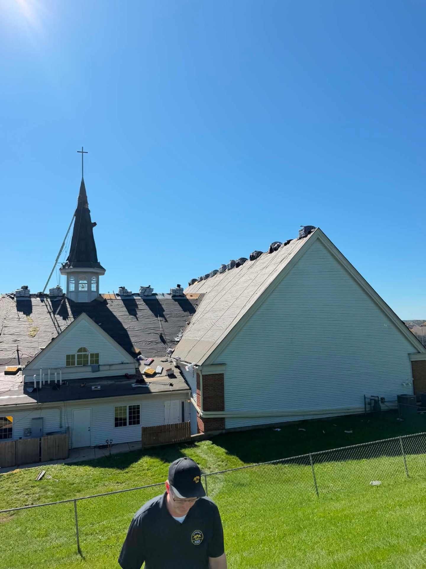 View across a chain-link fence toward a church roof covered in black underlayment, with shingle bundles along the ridges. A white steeple and cross rise behind the roof. A person in a dark shirt and cap stands in the foreground.
