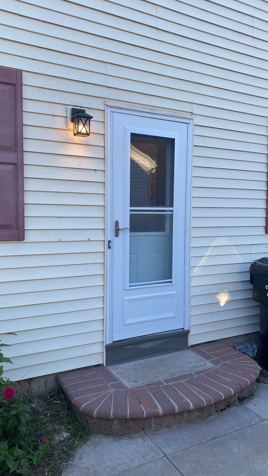 Finished white storm door with a screen, handle, and gray sill trim on cream siding, beside a lit wall lantern.