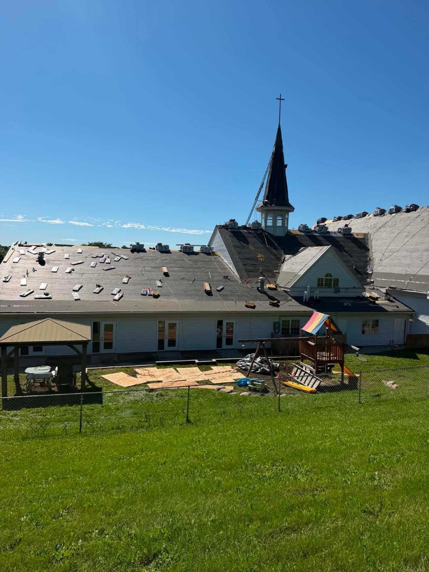 White church building with a steeple and cross. The roof is covered in black underlayment, with bundles of shingles staged along the ridge. A playground and gazebo sit in the fenced yard.