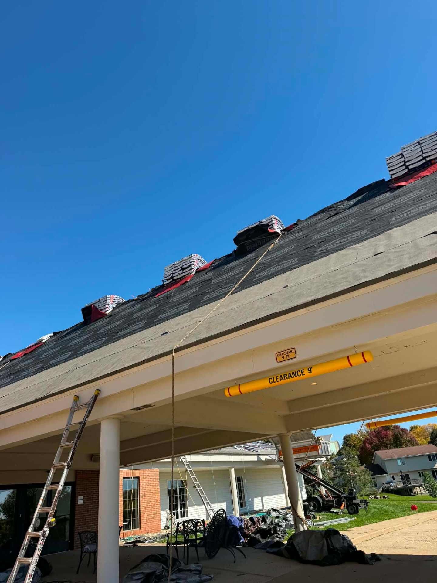 Low view of a roof edge covered in black underlayment, with bundles of shingles along the ridge, a ladder against a covered porch, and a yellow clearance bar.