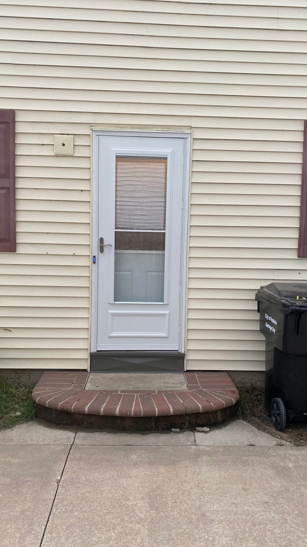 Finished white storm door with a screen and gray sill trim, centered on cream siding above a brick stoop.