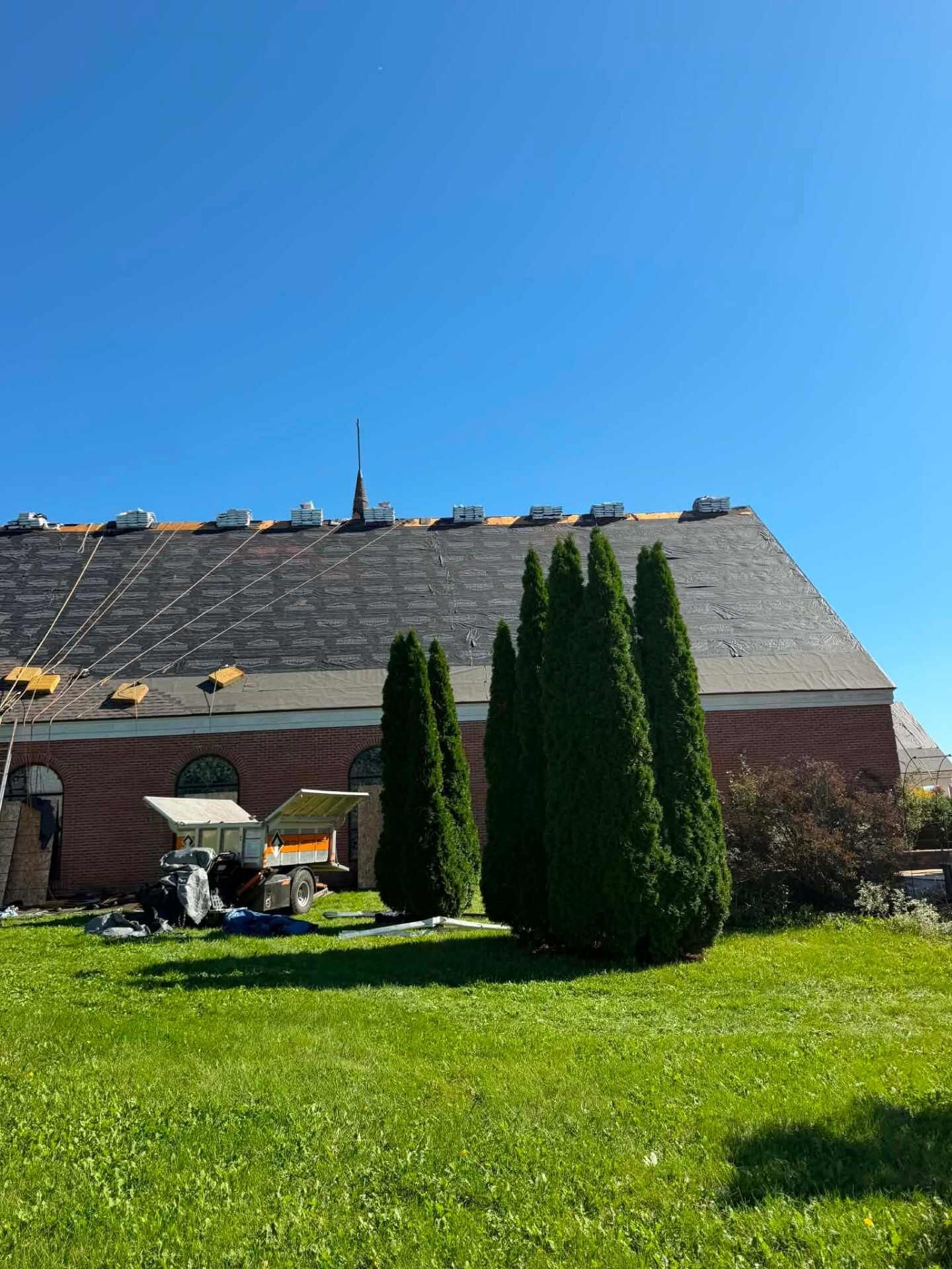 Brick wall under a steep roof covered in black underlayment, with shingle bundles along the ridge. Arborvitae stand in front of the wall, and a dump trailer and torn-off roofing debris sit on the grass.