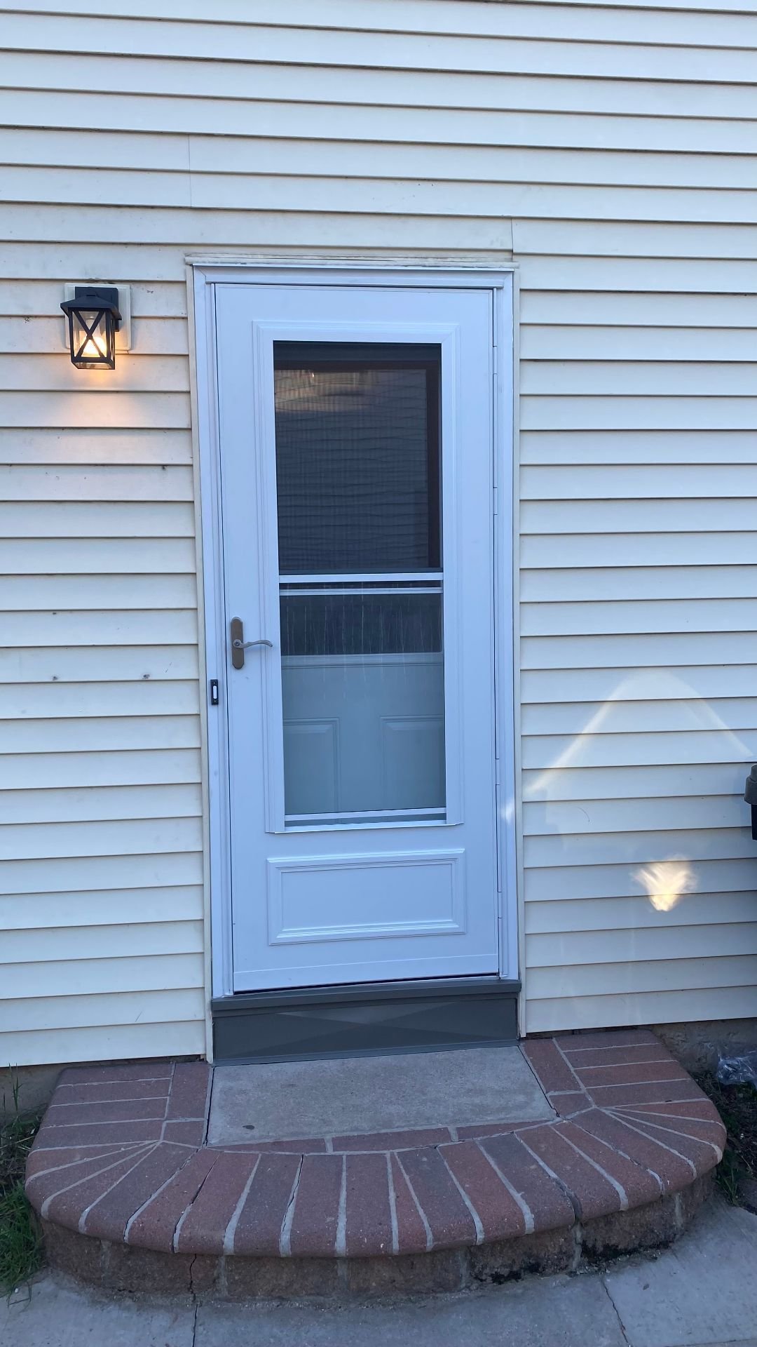 Close view of a finished white storm door with a screen, handle, and gray sill trim on cream siding.