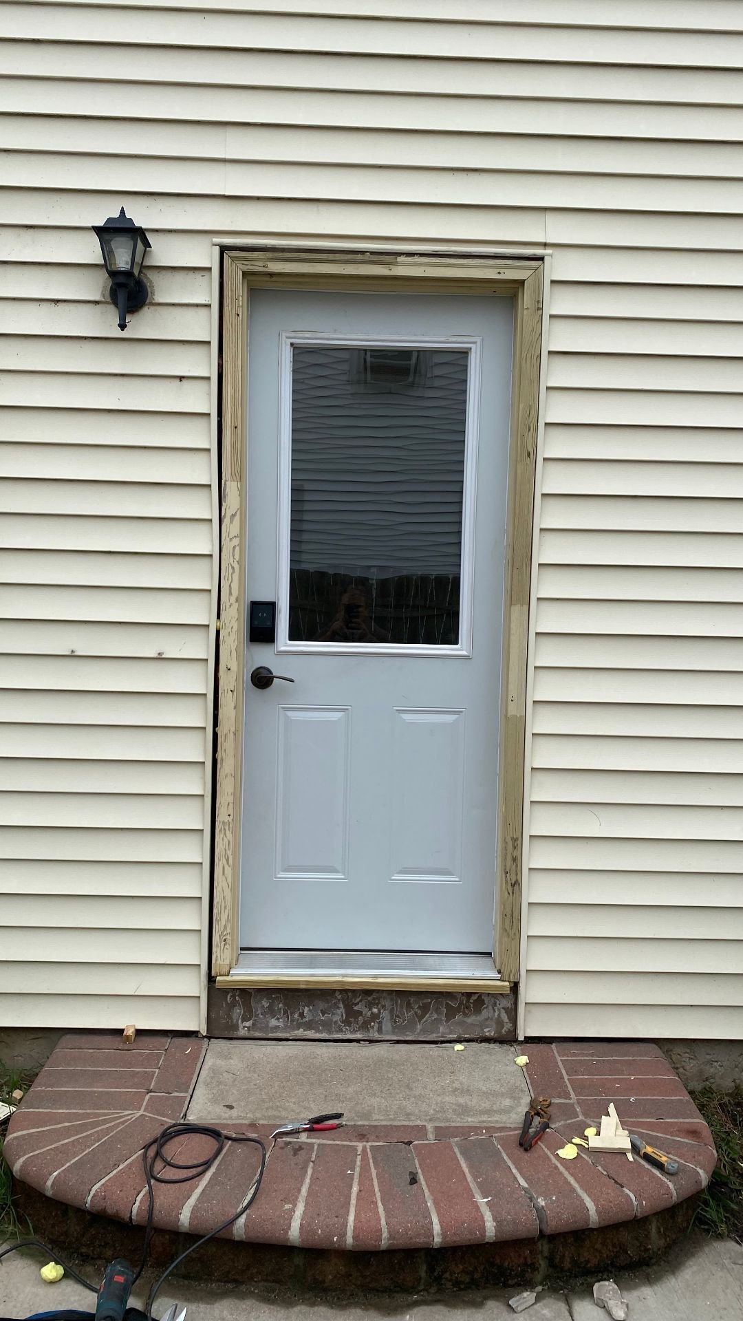 White entry door set in an unfinished wood jamb on cream siding, with tools on a brick stoop during storm door installation.