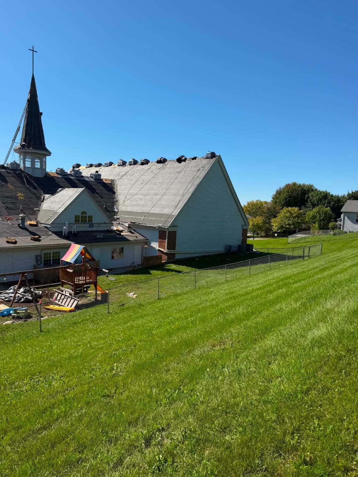 White church with a steeple and cross. Black underlayment covers part of the roof, and a light underlayment covers the large gable. A playground with a colorful canopy sits behind a chain-link fence.