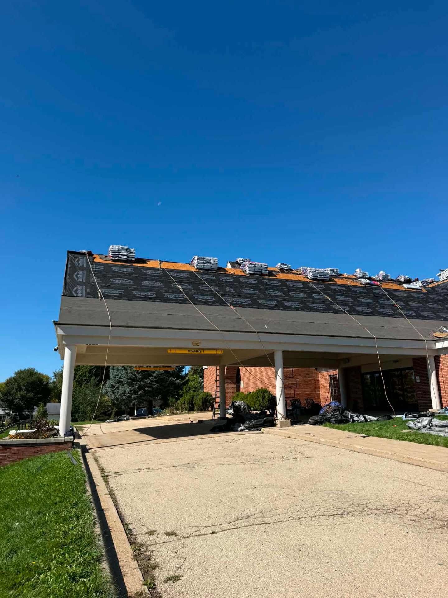 Covered porch with white columns and a yellow clearance bar. Black underlayment covers the roof above, with shingle bundles along the ridge. Torn-off roofing debris is piled on the concrete under the porch.