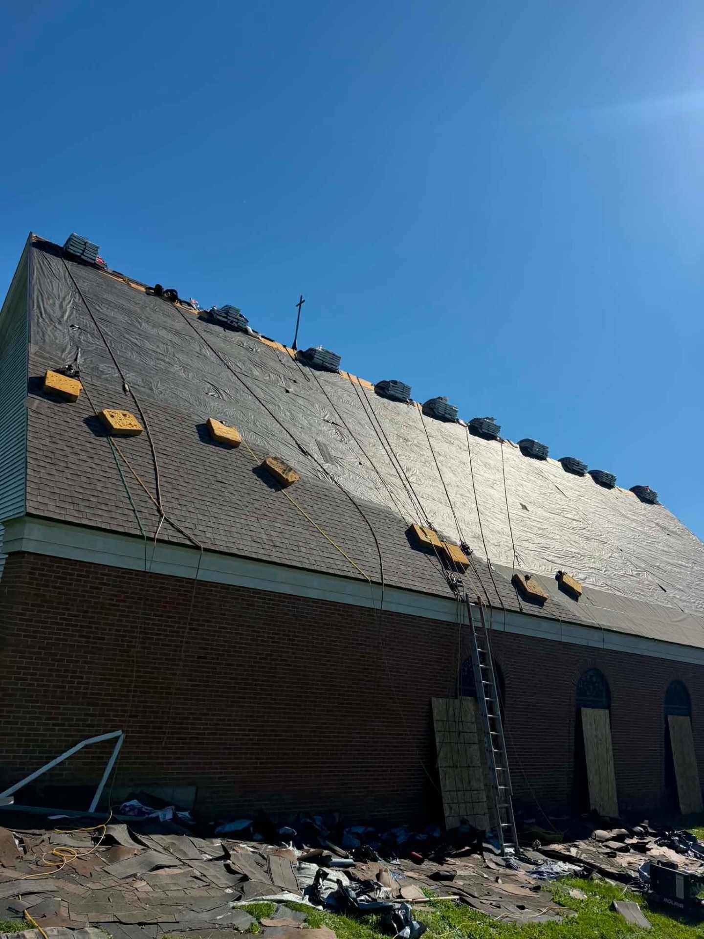 Steep roof on a brick wall, partly covered with dark shingles and partly with light underlayment. Torn-off roofing debris is on the ground, plywood covers arched openings, and a ladder leans on the wall.