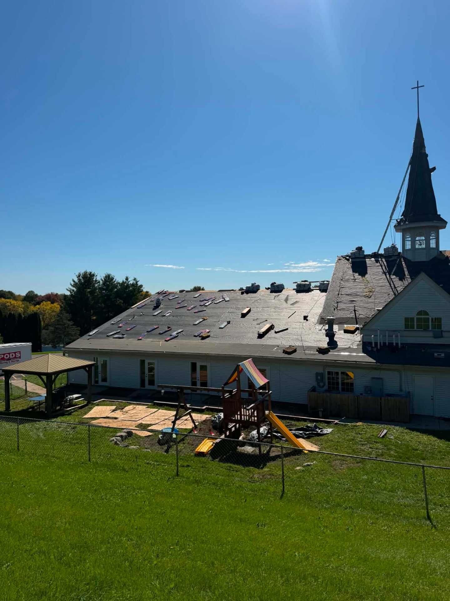 Side of a white church with a steeple and cross. Black underlayment covers the roof, with shingle bundles near the ridge. A playground and gazebo are in the yard.