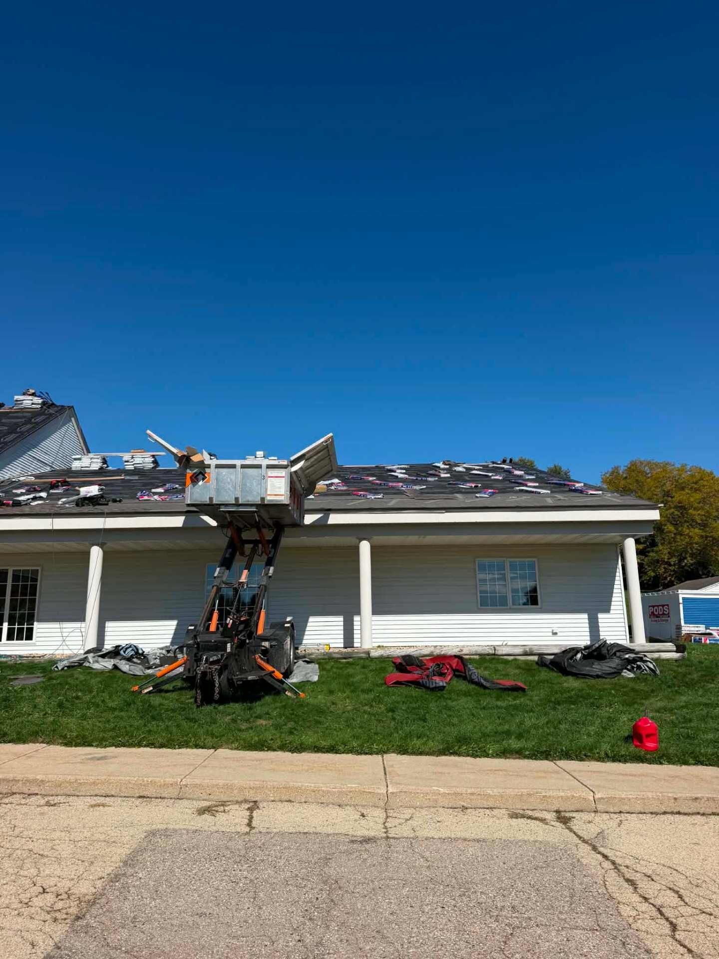 White building with a covered porch. A lift is raised to the eave, and bundles of shingles are staged across black underlayment on the roof.