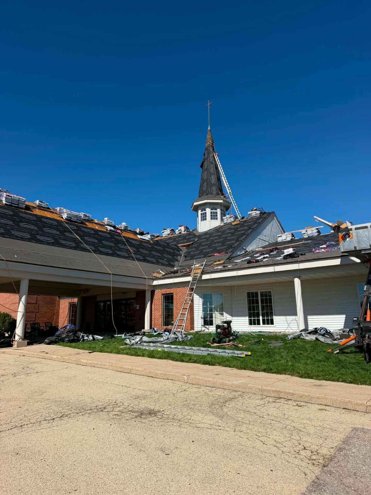 Church front with a steeple and cross. The roof has black underlayment and bundles of shingles along the ridges, with ladders and a lift at the building.