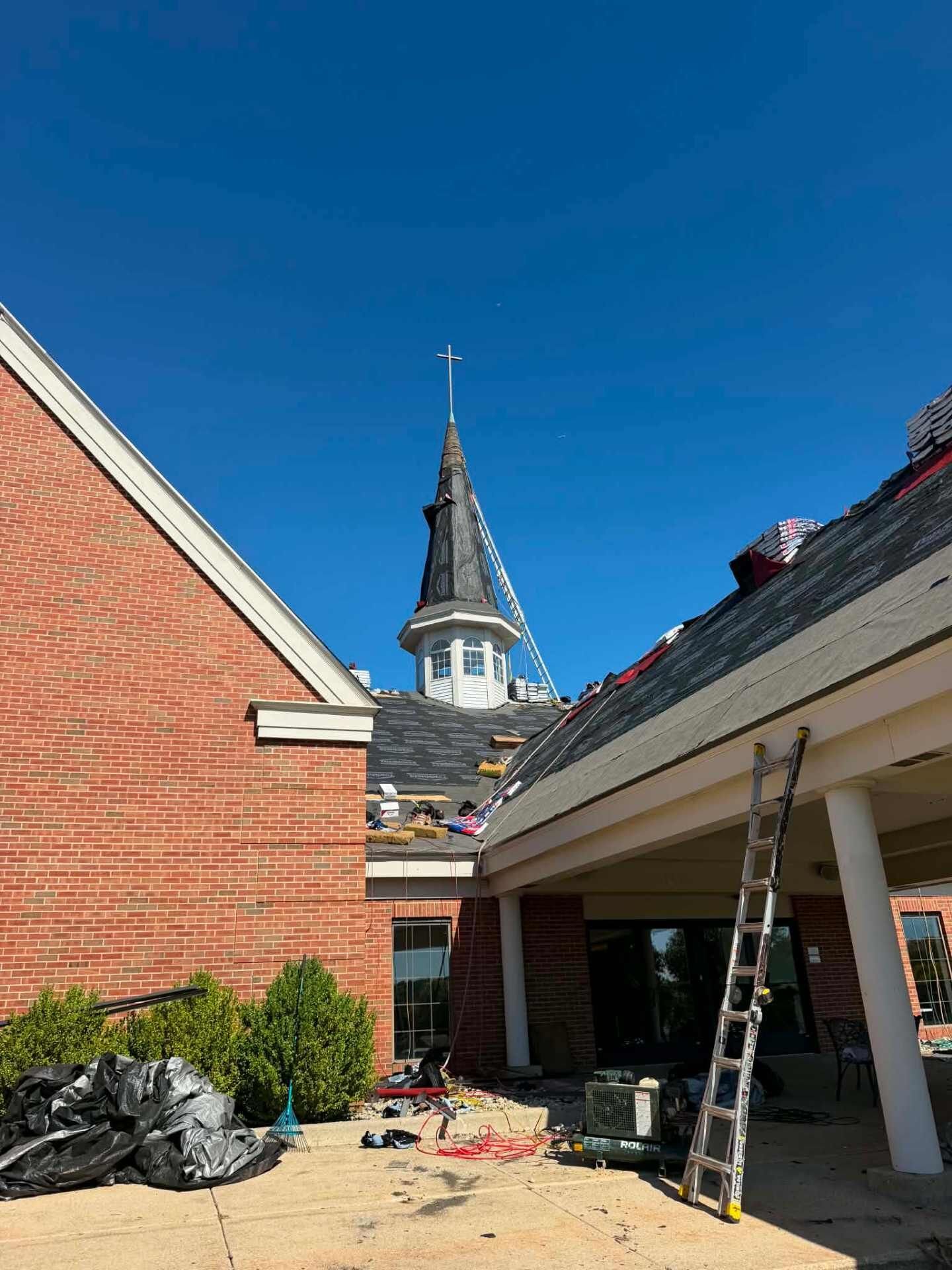 Brick church wall and covered porch. The steeple is wrapped in black with a cross at the top. A ladder stands on the porch, and torn-off roofing debris is piled on the concrete.