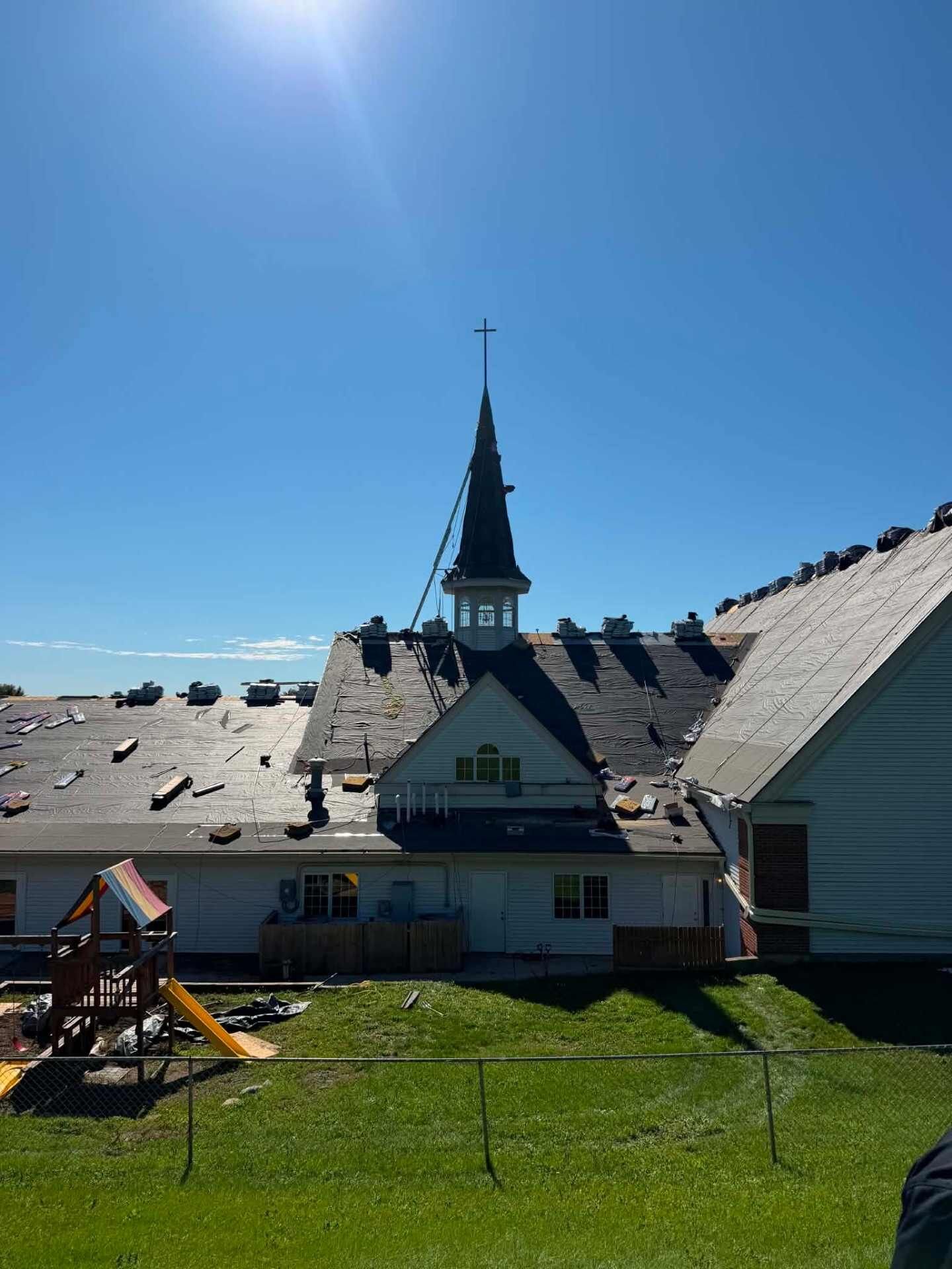 Church roof covered in black underlayment, with bundles of shingles staged across it and a white steeple and cross above. A playground with a yellow slide sits in the fenced yard.