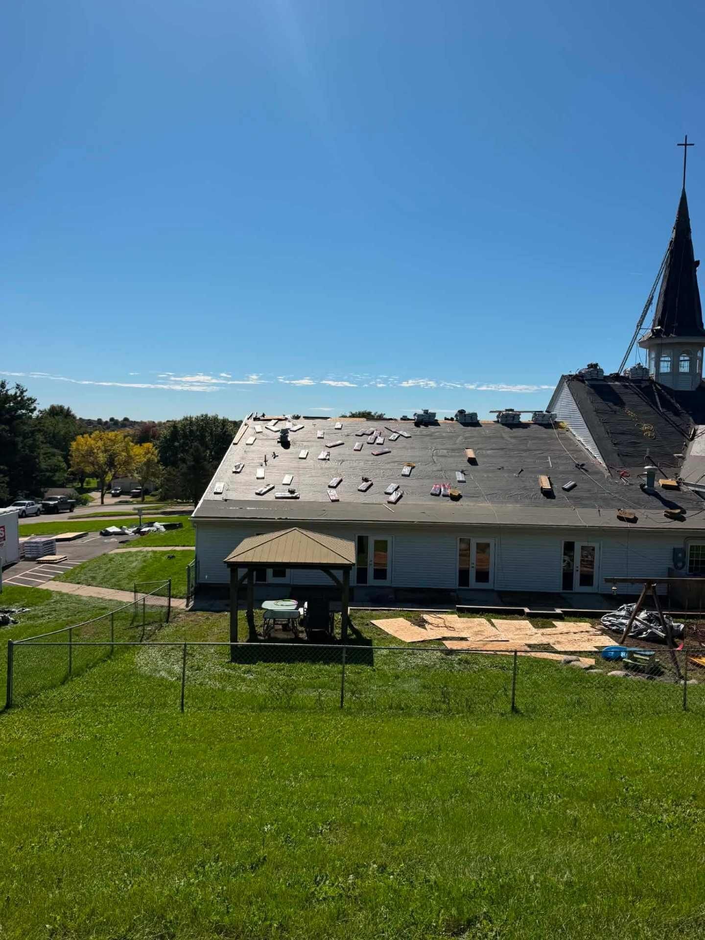 White church with a steeple and cross. Black underlayment covers the roof, with shingle bundles and loose pieces scattered across it. A gazebo and plywood sheets sit in the fenced yard.