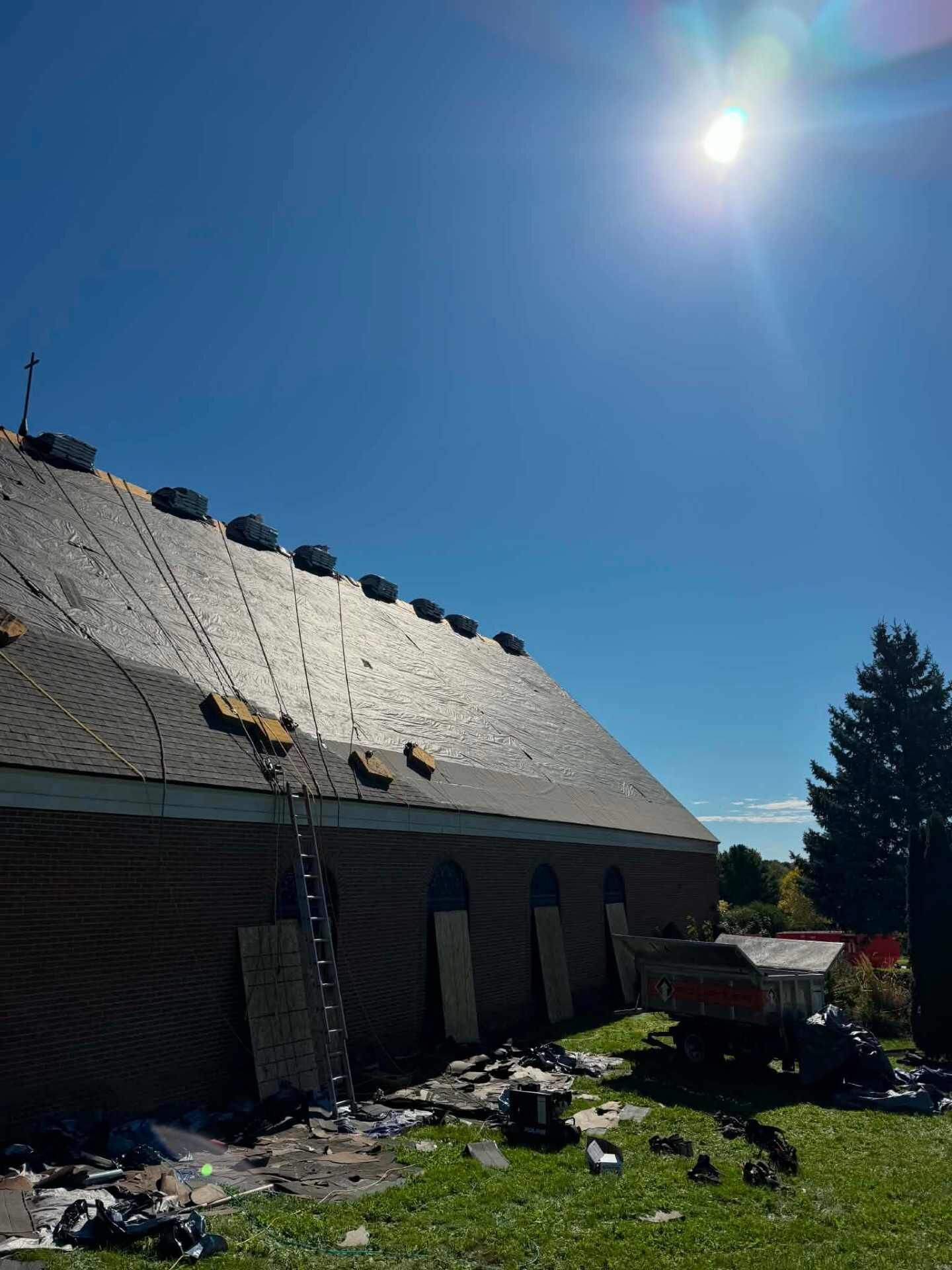 Brick wall under a steep roof that is partly dark shingles and partly light underlayment, with vents along the ridge. A ladder leans on the wall, torn-off roofing debris covers the grass, and a dump trailer sits nearby.
