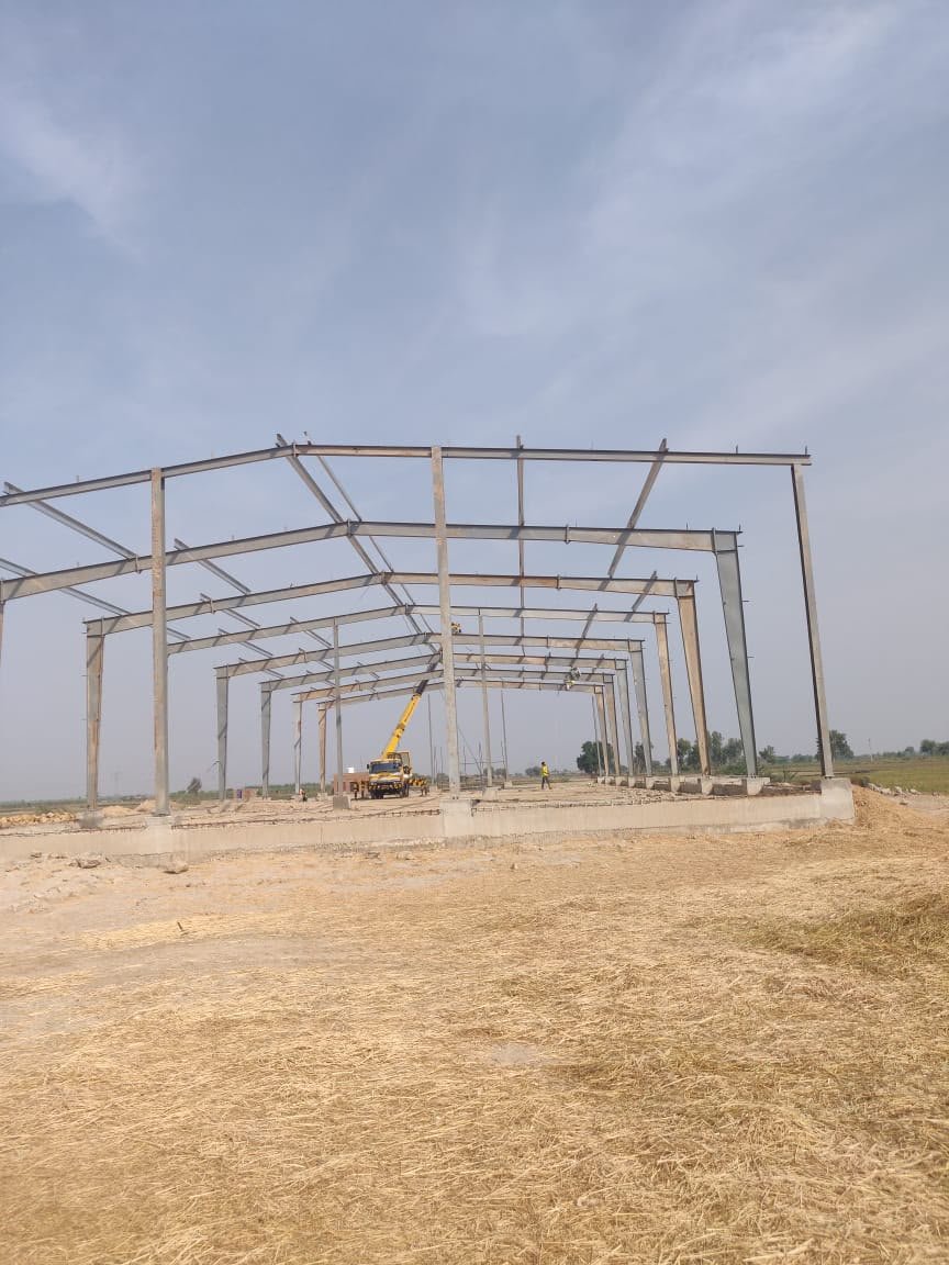 Multi-bay steel portal frame shed under erection on a rural site, with a yellow crane inside the frame line