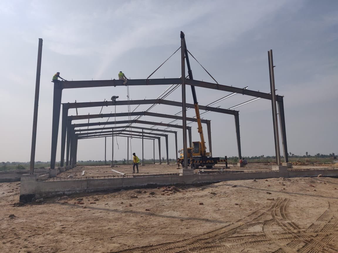 Workers bolting a steel beam on a portal frame while a yellow crane lifts the next rafter into place