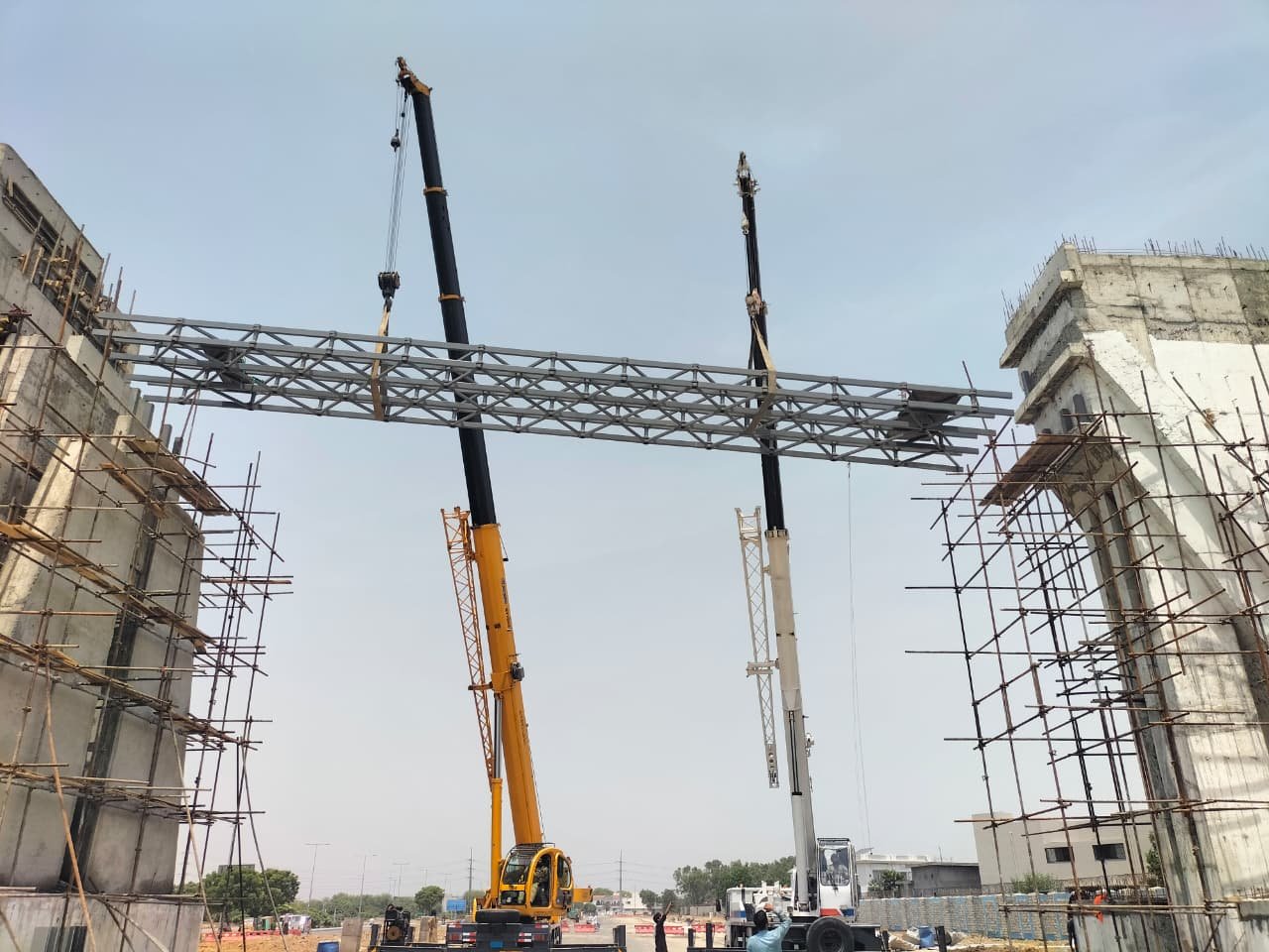 Grid shell gateway frame between concrete pylons with cranes and workers on site