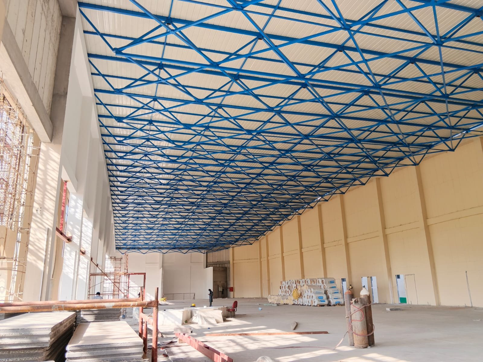 Wide hall under a blue steel roof lattice, with stacked panels and gas cylinders on the floor