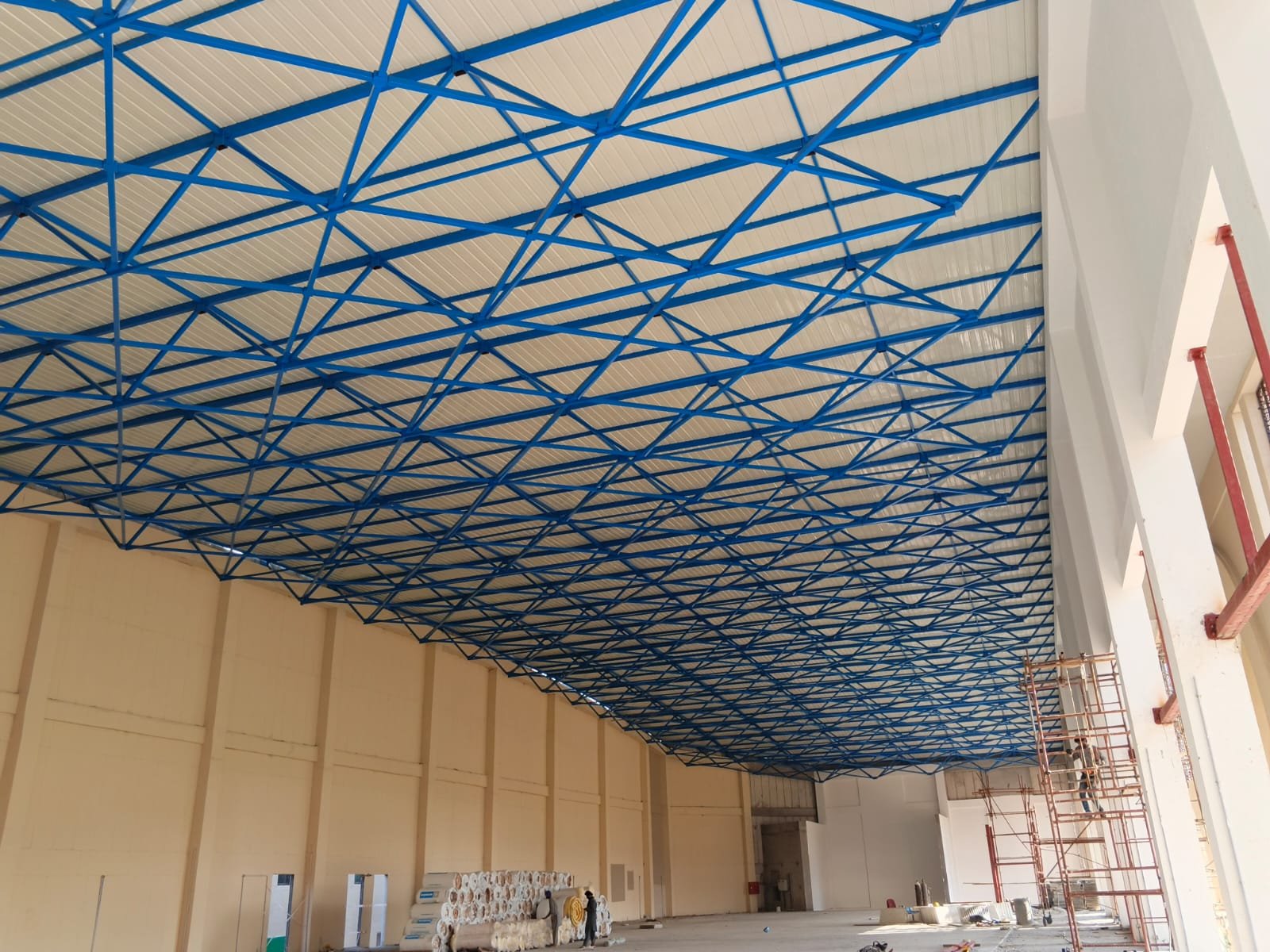 Blue steel roof lattice under construction inside a large hall, with scaffolding and insulation rolls