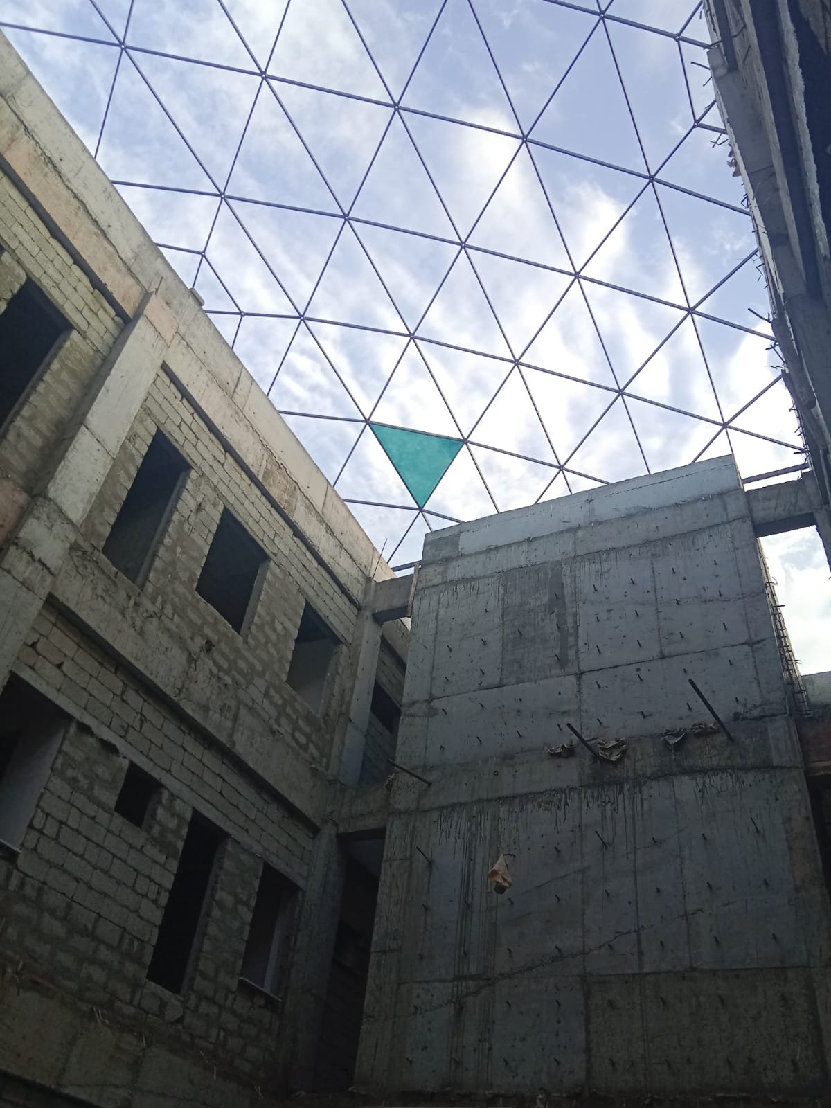 View upward through a geometric steel roof lattice between concrete walls, with one teal panel