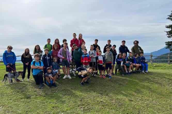 Group of children and teenagers of various ages gathered together in a meadow with mountains and lake in the background
