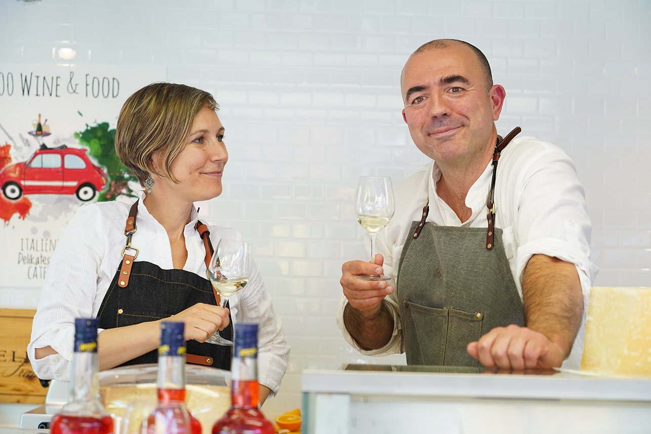 Michele und Alexandra, Gründer von 500 Wine & Food, lachend an ihrem italienischen Marktstand mit Wein und Parmesan auf einem Wochenmarkt