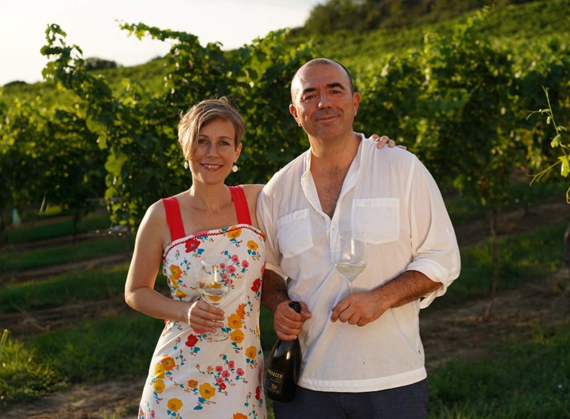 Michele und Alexandra am Wochenmarkt-Stand in Niederösterreich — Michele mit einer Flasche italienischem Wein, Alexandra mit einem Stück Parmesan aus Italien