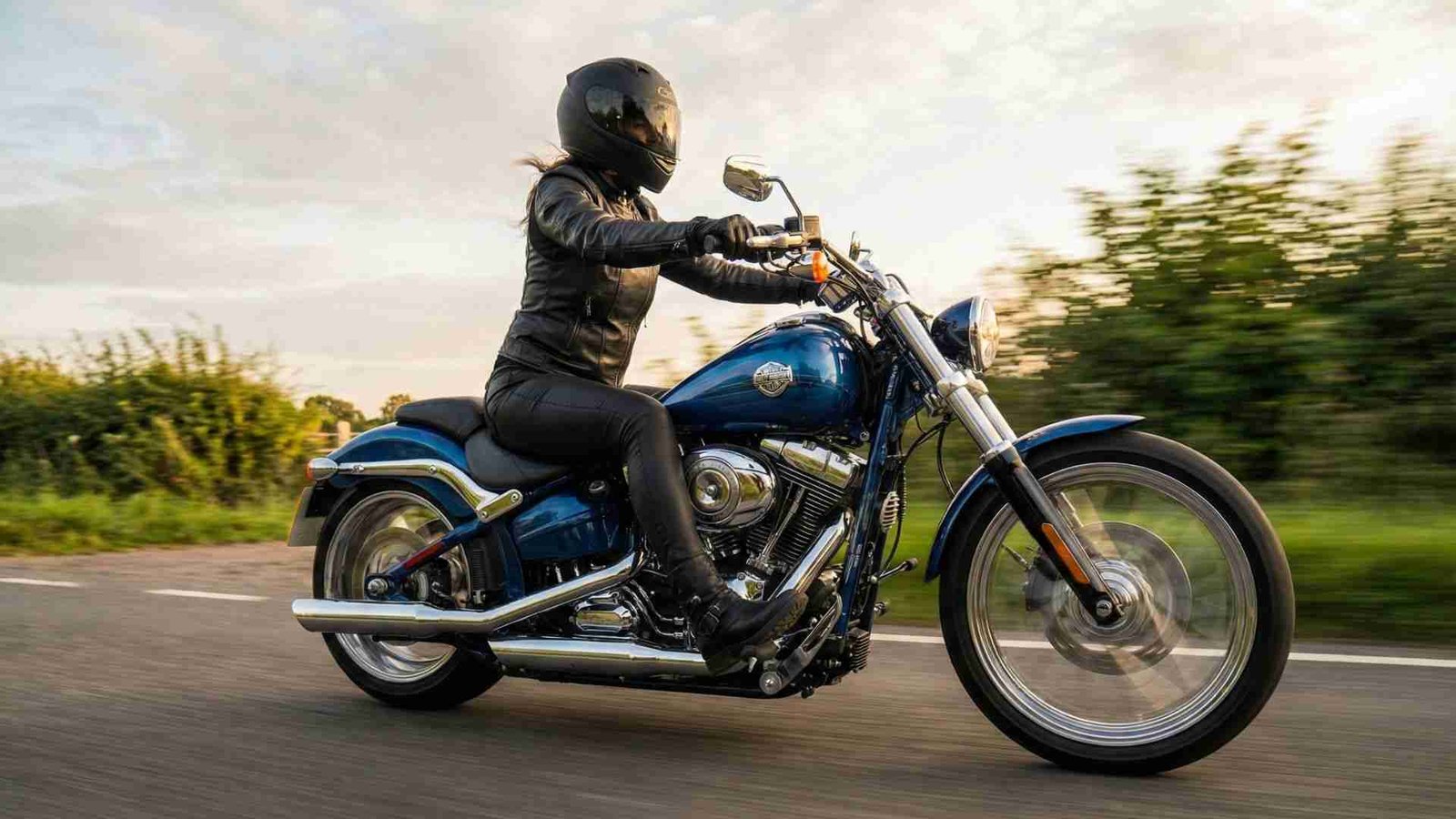 Mulher motociclista com capacete e jaqueta de couro pilotando uma Harley Davidson azul em alta velocidade em estrada rural ao pôr do sol