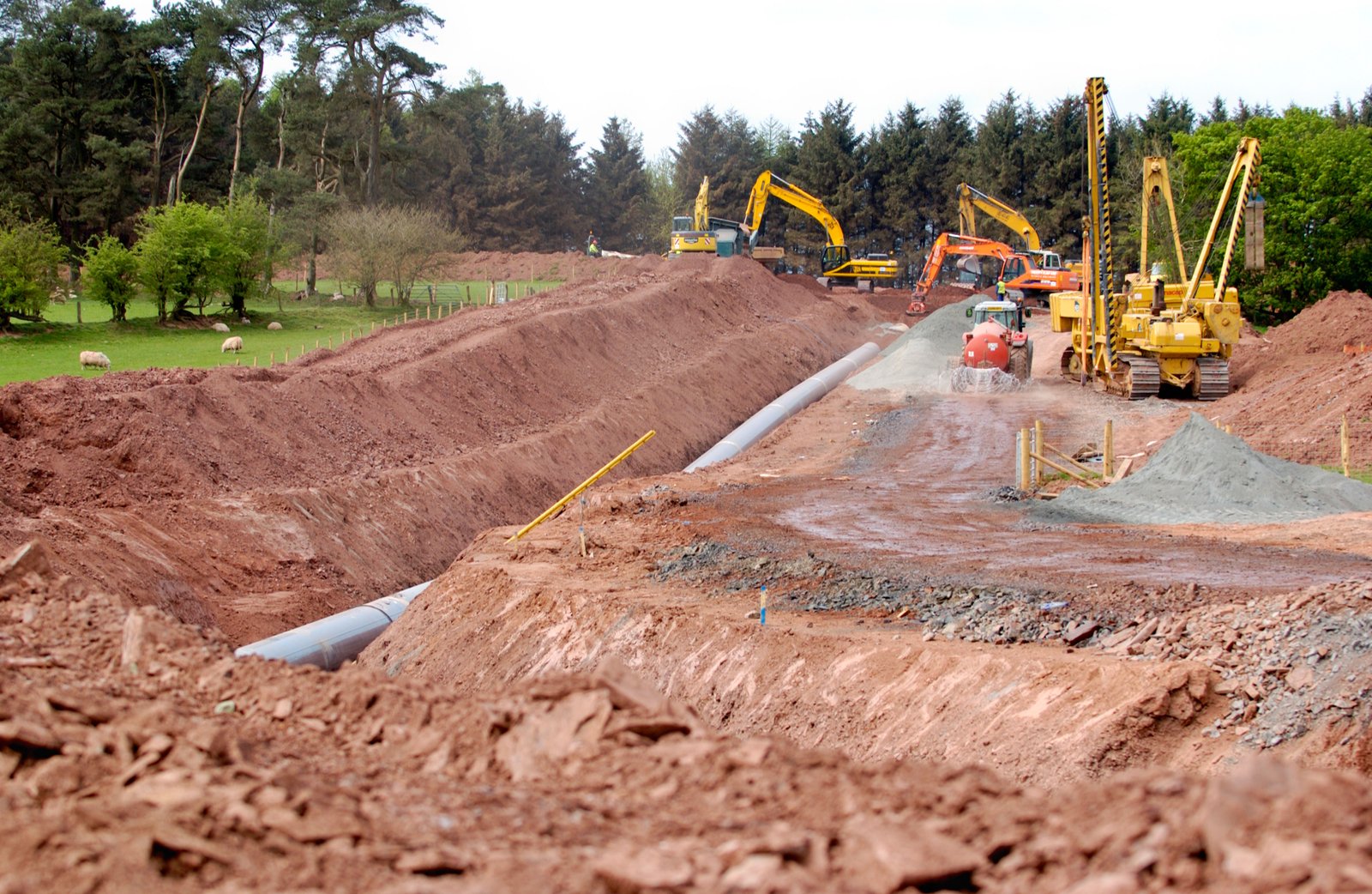 UK pipeline construction corridor with excavators and red earth working strip through woodland edge
