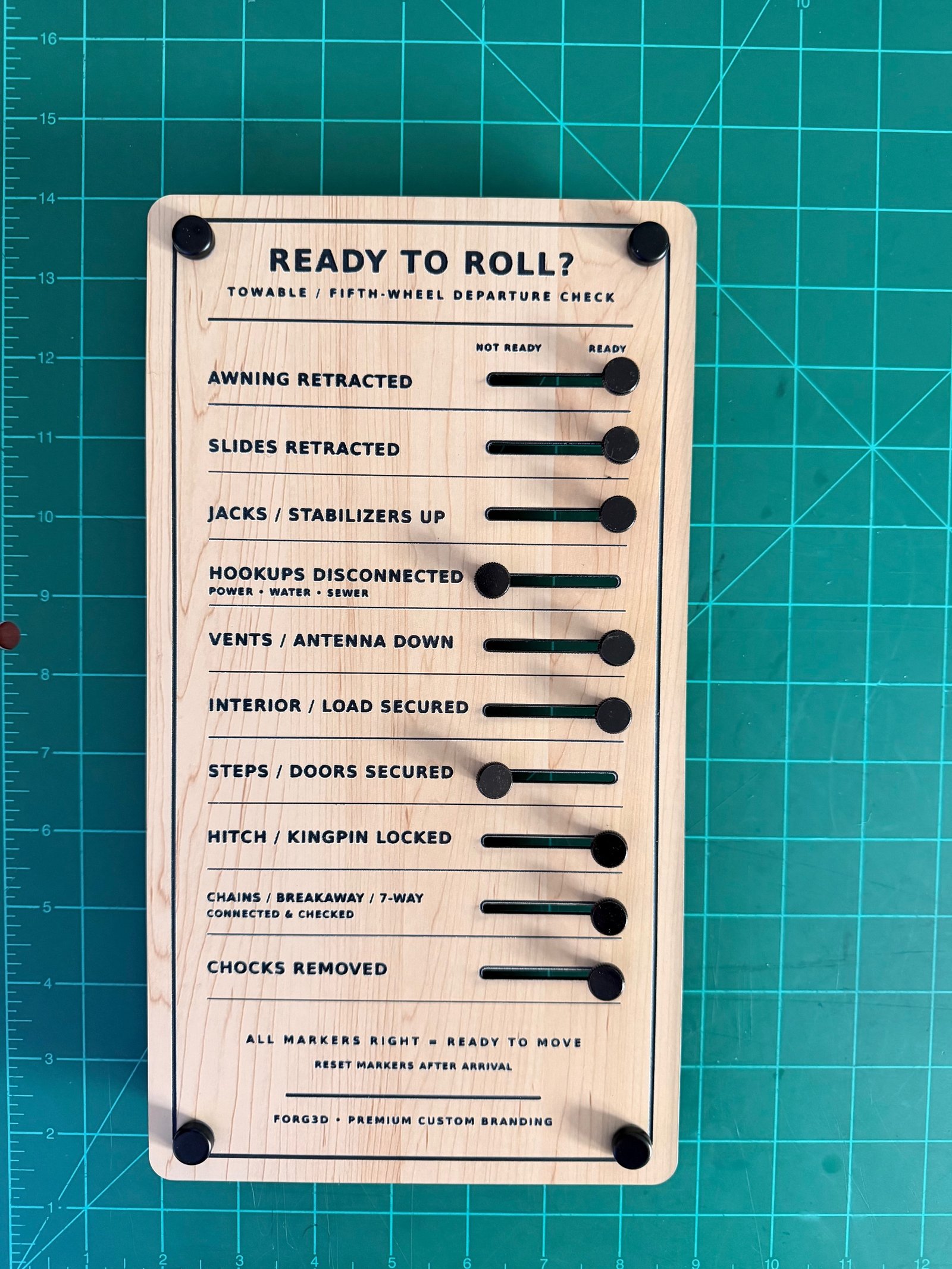 Portrait of a light wood-grain FORG3D Ready to Roll towable and fifth-wheel departure checklist board on a green cutting mat, with black sliding toggles and corner standoffs