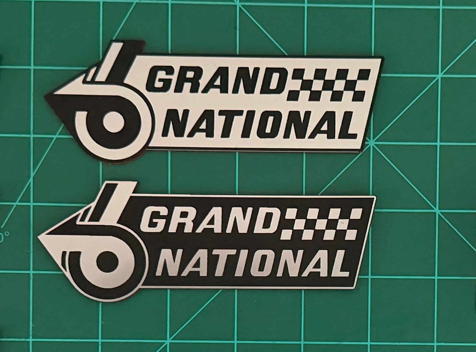 White and black Grand National acrylic badge pair photographed on a green cutting mat