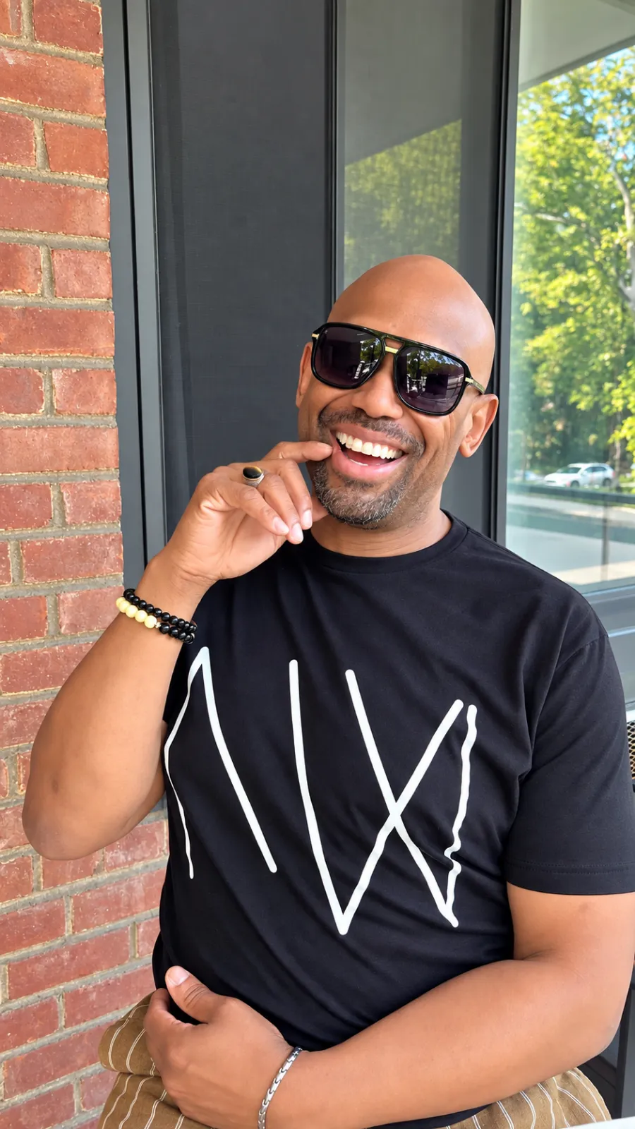 Corey Wesley laughing outdoors in sunglasses, seated beside a brick wall