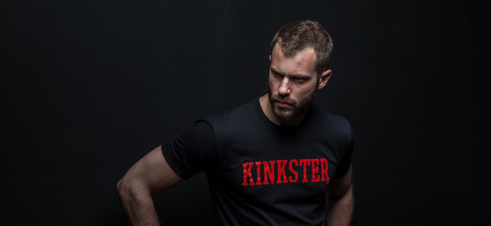 Bearded model in a black Kinkster T-shirt, looking down against a dark studio wall