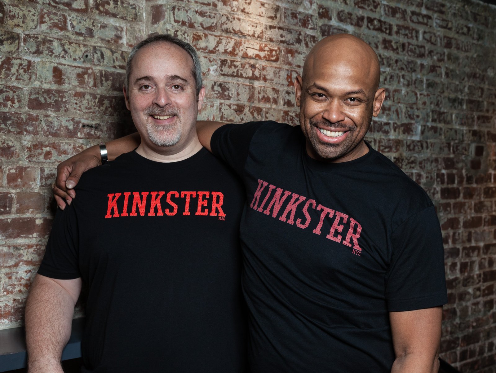 Two men in black Kinkster T-shirts standing together against a brick wall