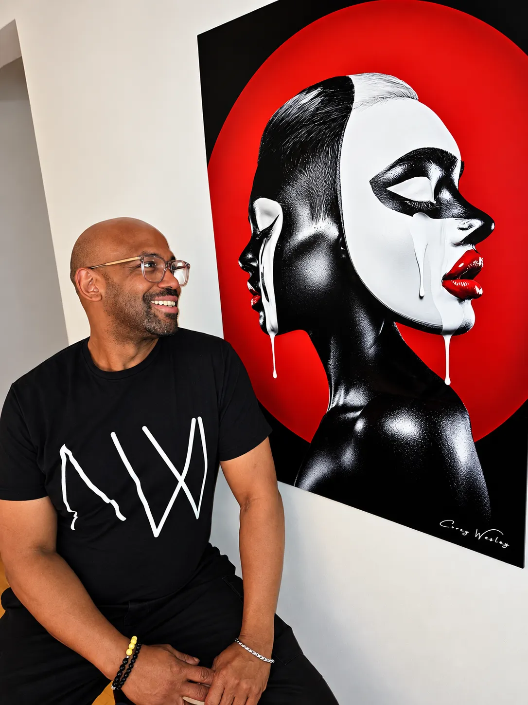 Corey Wesley seated beside a large black-and-red portrait artwork, wearing glasses and a black T-shirt