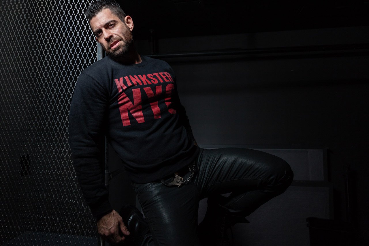 Model in a black Kinkster NYC sweatshirt and leather pants leaning against a chain-link fence