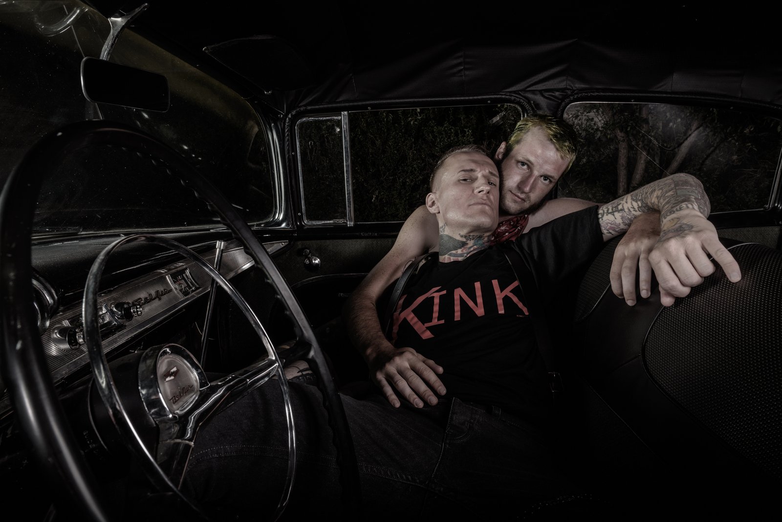Two men in a vintage car at night, one wearing a black Kink shirt