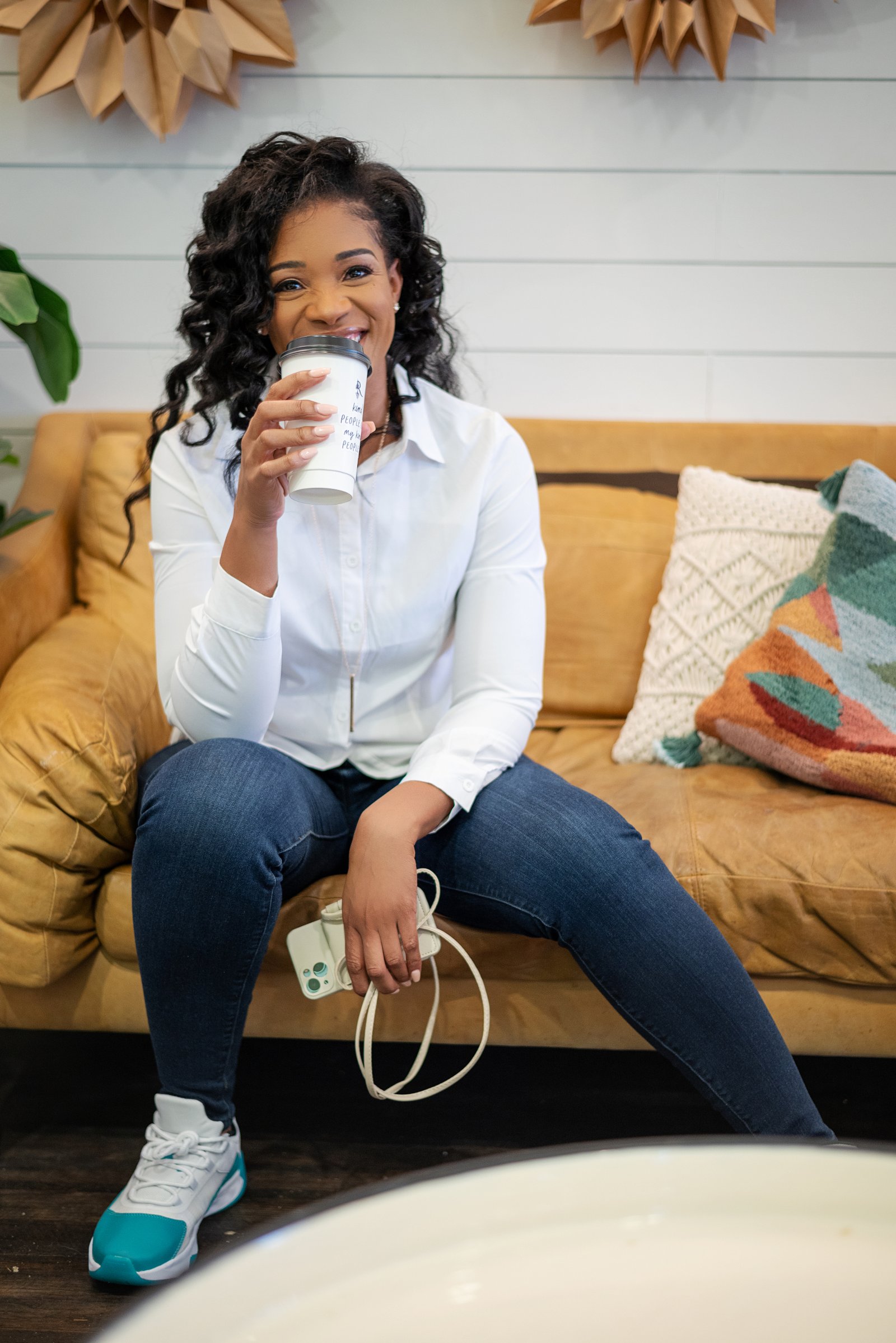 Professional headshot of Thameka Marisa sitting on a mustard yellow couch, wearing a white button-up shirt and dark jeans, holding a white coffee cup, smiling warmly at the camera