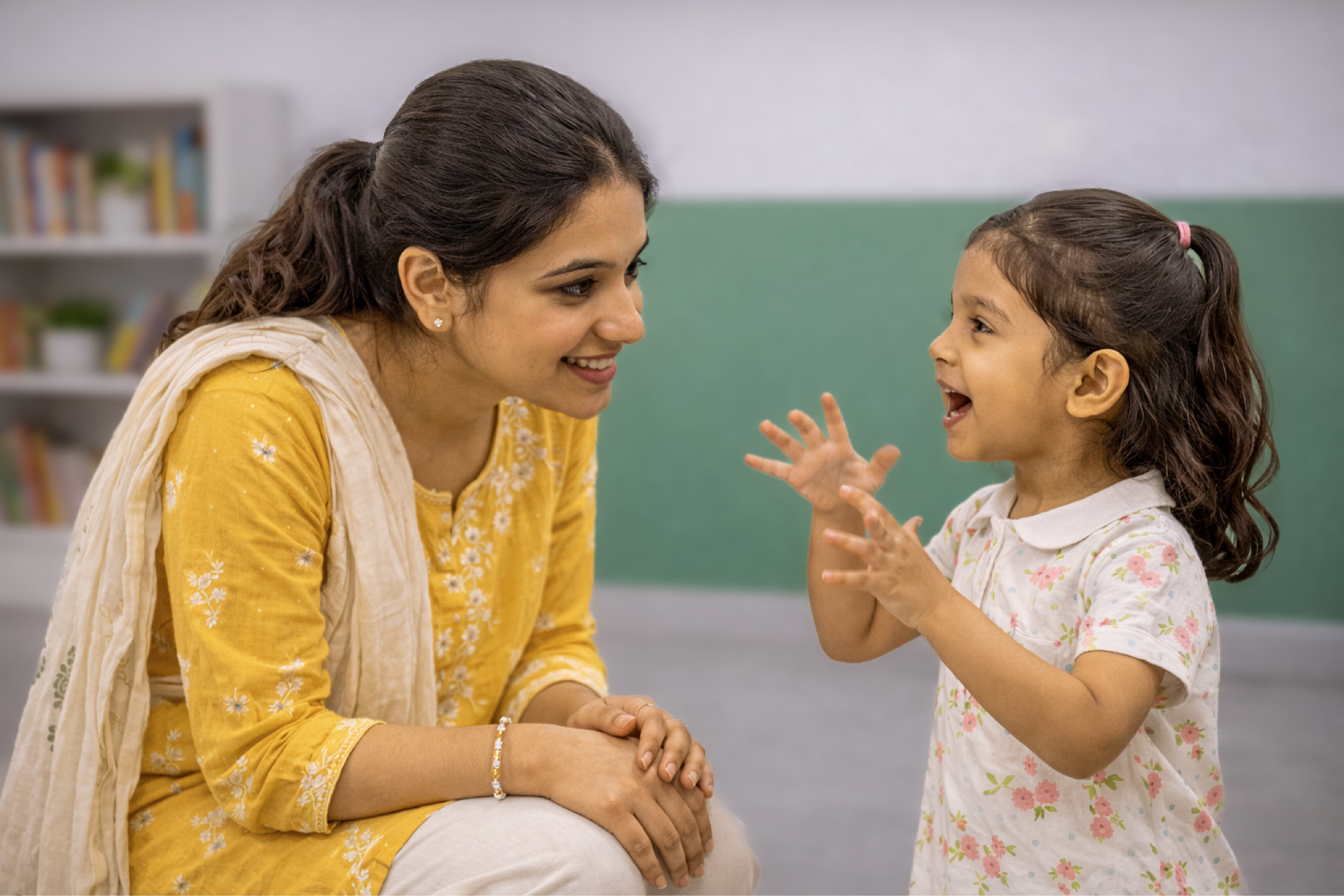 Woman in yellow traditional dress with beige shawl smiling warmly at young girl in classroom setting