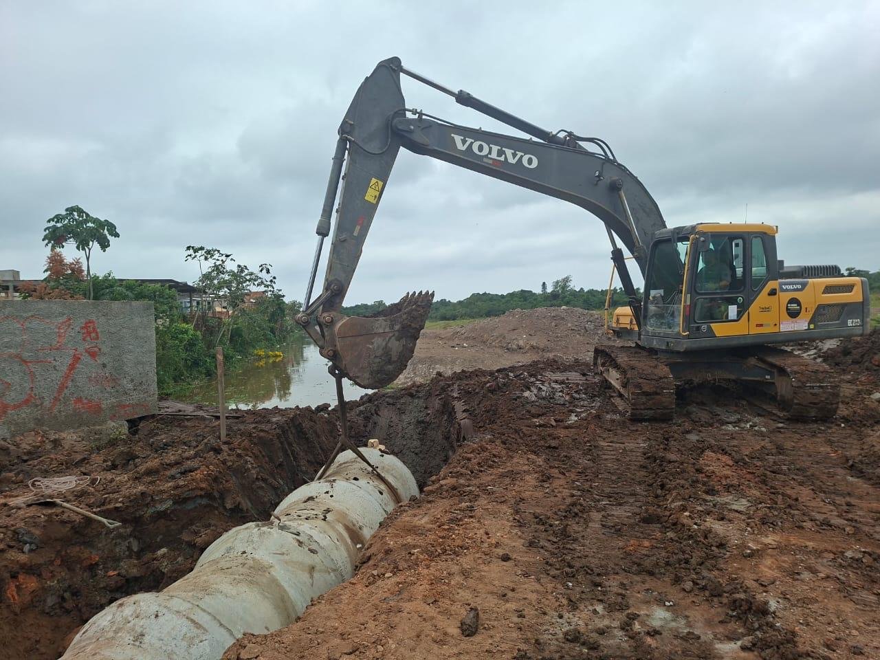 Escavadeira posicionando tubulação de concreto em vala de drenagem