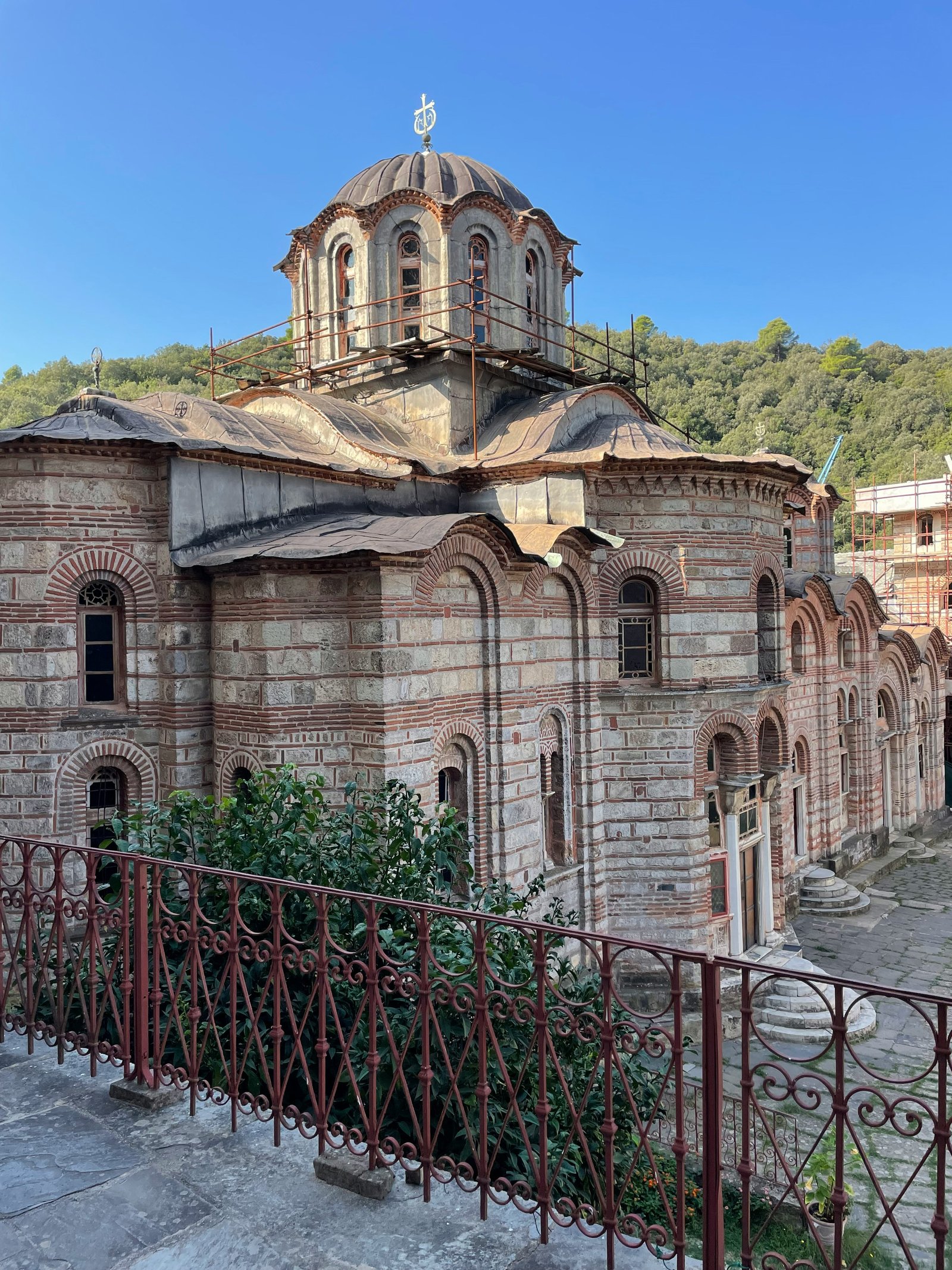 Byzantine monastery with ornate dome, red brick arches, stone columns, iron railing, forest background, clear blue sky