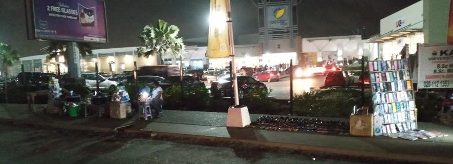 Wide nighttime view of roadside trading and illuminated commercial buildings in the Accra Mall Enclave.