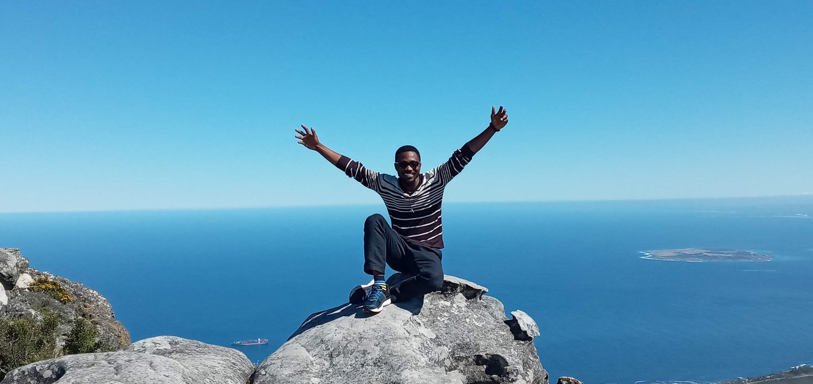 Joe Quarcoo seated on a rock at the summit of Table Mountain with the Atlantic coastline behind him.