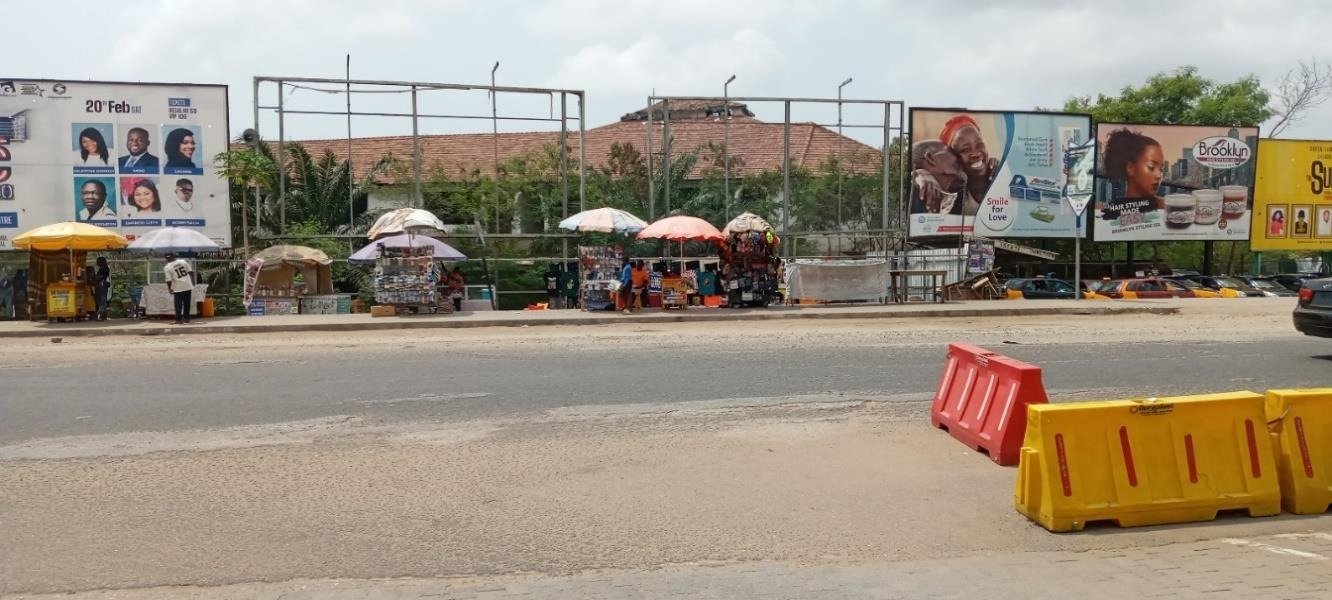 Roadside vendors trading beneath billboards in the Accra Mall Enclave during the day.