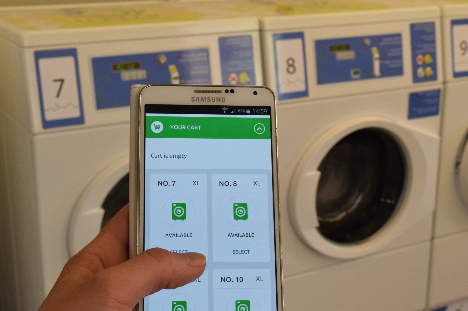 A hand holding a phone with a laundry-machine cart on screen in front of commercial washers