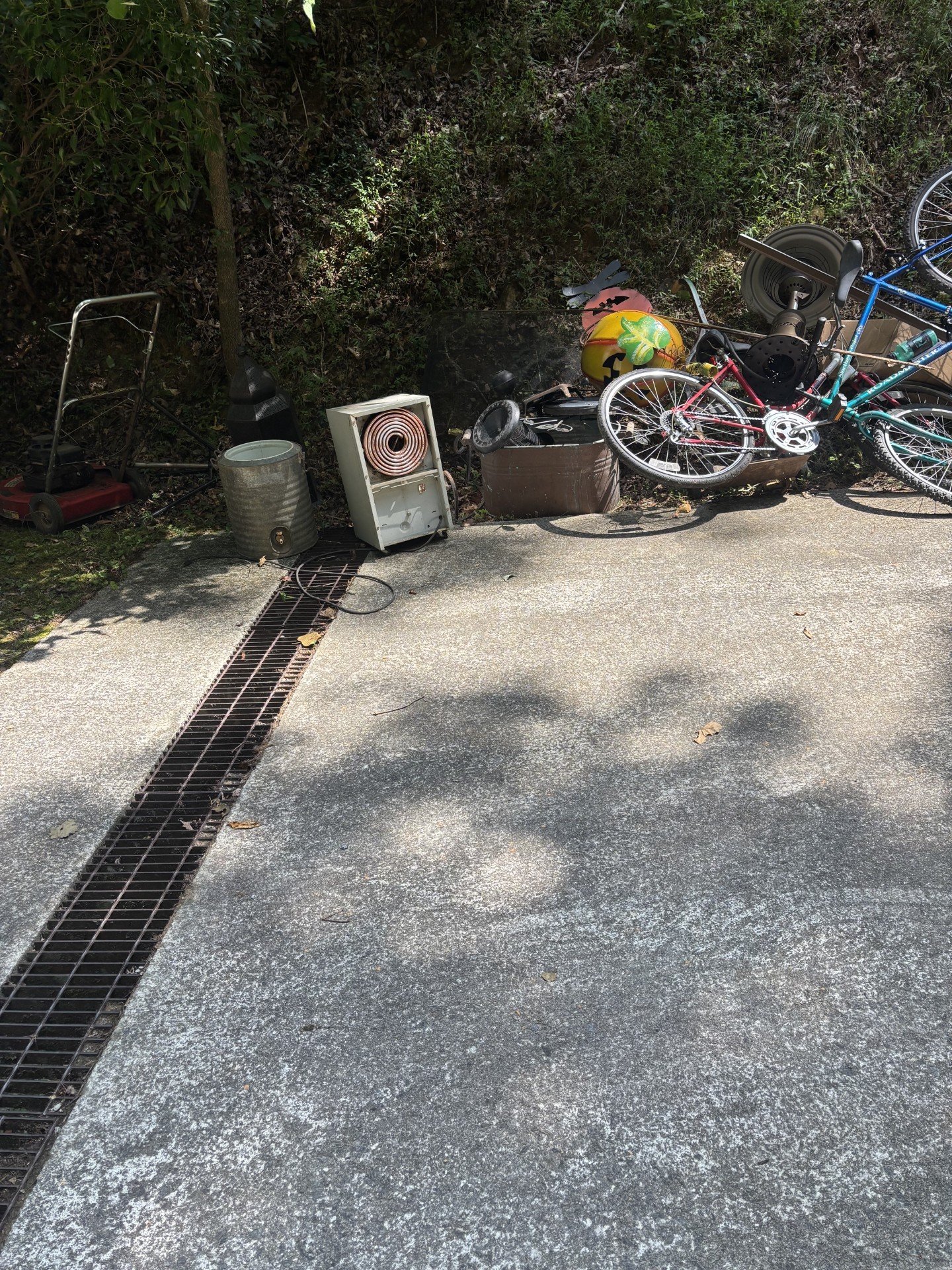 Driveway with junk piled up including ladder, metal drum, dehumidifier, bikes, helmets, and various items before removal