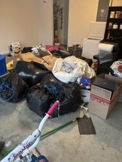 Cluttered garage with black trash bags, boxes, and miscellaneous items before cleanout